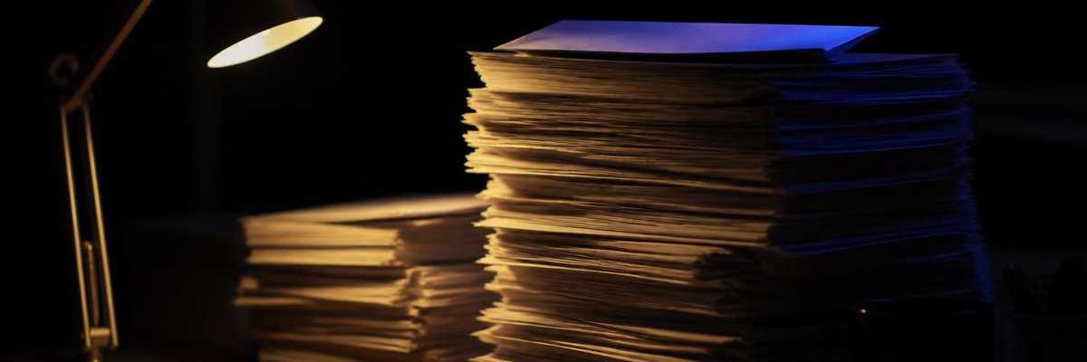 Cinematic night still life of tall paper stacks on a desk, a single envelope on the highest stack edged in faint indigo light